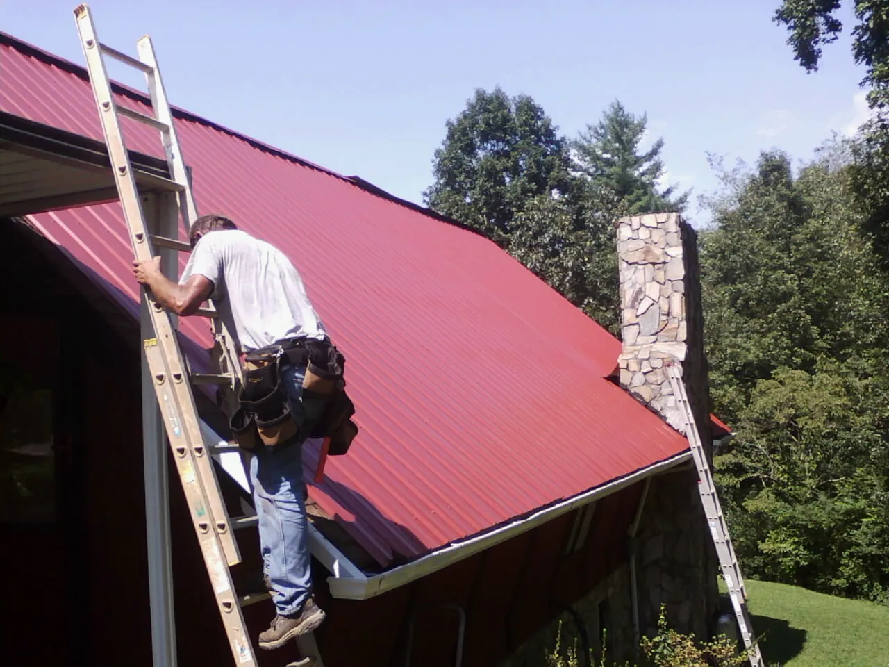 Roofer on a ladder beside a red metal roof and stone chimney — Soffit & Fascia Repair by Roofing Brothers in Deersville